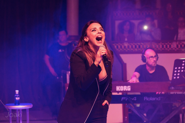 Frau singt in ein Mikrofon auf der Buhne mit einem Pianisten und Publikum im Hintergrund, eine Flasche auf einem Hocker zu ihrer Linken.