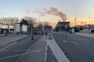 Eine Stadtstraße mit Verkehrskegeln, einem Bus, Laternen, Bäumen, Gebäuden, Kränen und Rauch, der aus einem entfernten Gebäude aufsteigt, unter einem klaren Himmel.