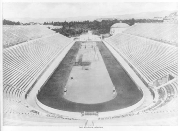 Altes Schwarz-Weiß-Foto des Olympischen Stadions in Athen, Griechenland, mit Sitzreihen, Bäumen, Gebäuden und Bergen im Hintergrund sowie Text am unteren Bildrand.