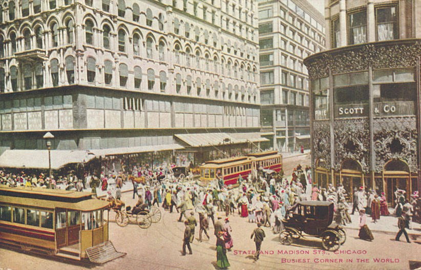 Altes Schwarz-Weiß-Foto einer belebten Stadtstraße mit Fußgängern, Pferdewagen und Laternen, Gebäuden mit Fenstern im Hintergrund und Text am unteren Rand.