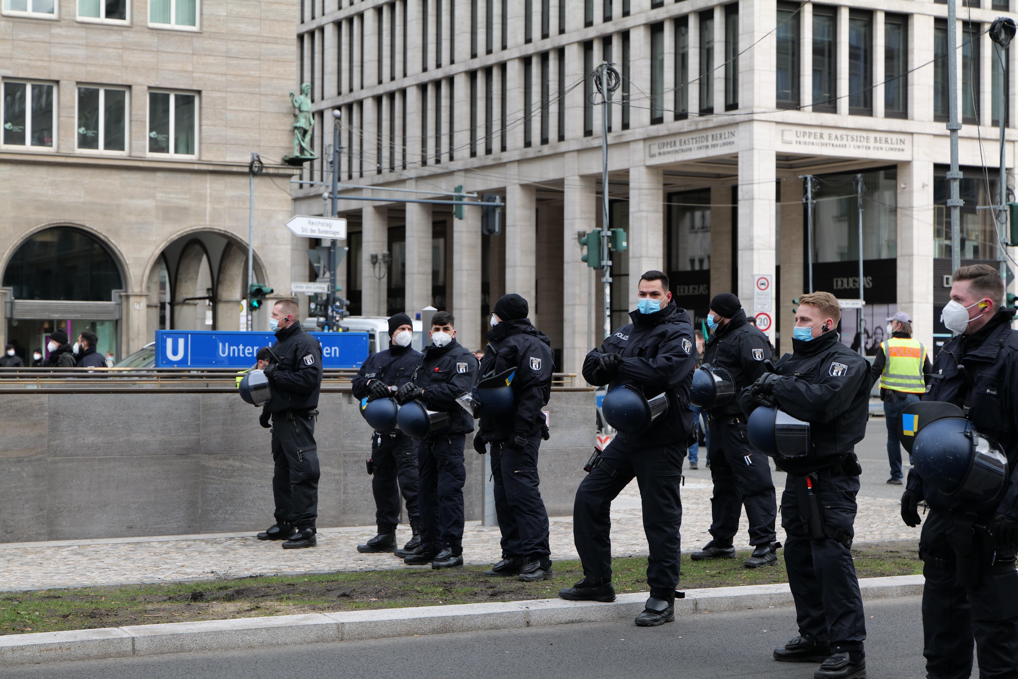 Polizisten in schwarzen Uniformen und Masken vor einem Berliner Gebäude mit Glasfenstern, Pfeilern und umgebenden urbanen Elementen.