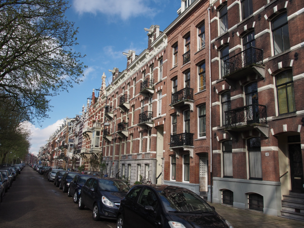 Eine Reihe geparkter Autos entlang einer Baumreihe in Amsterdam, mit Fenstern, Geländern und Stufen versehene Gebäude im Hintergrund unter einem bewölkten Himmel.