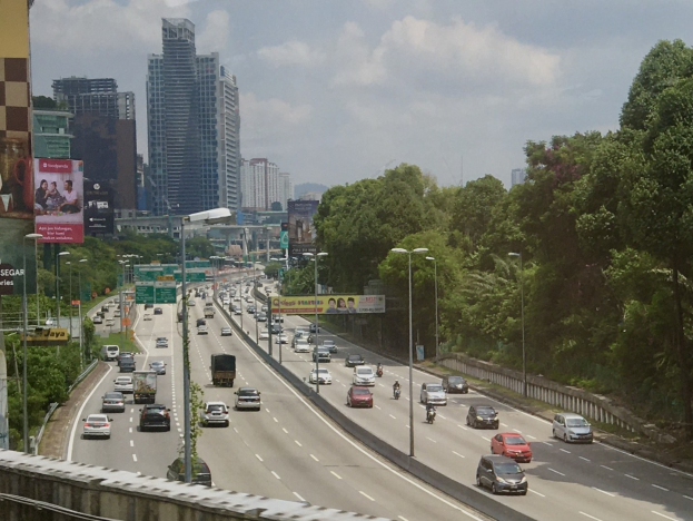 Eine vielbefahrene Autobahn mit hohen Gebäuden, Straßenlaternen, Lichtern, Werbetafeln, Bäumen und bewölktem Himmel.