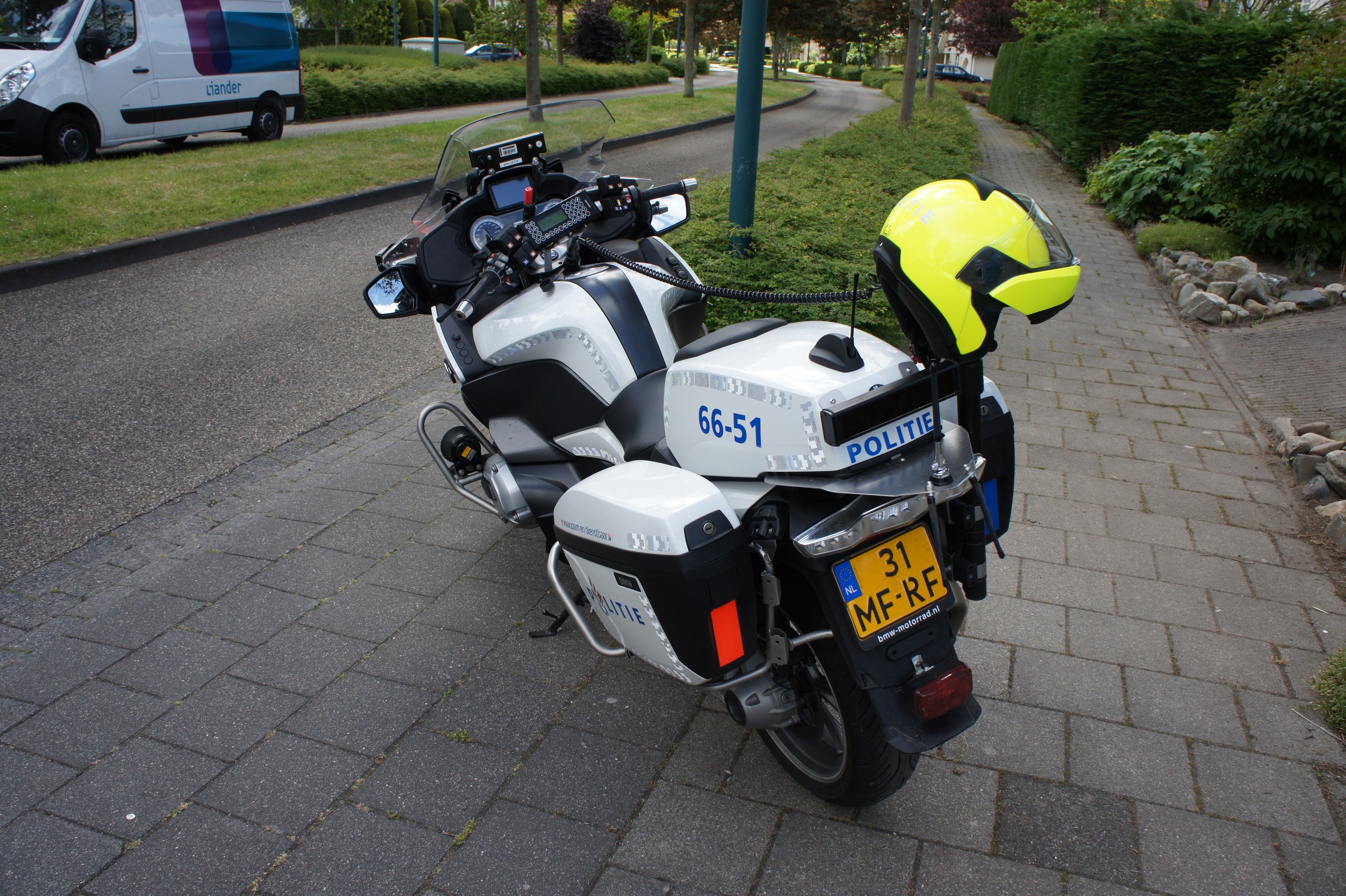 Ein Polizeimotorrad am Straßenrand geparkt mit einem Helm darauf, umgeben von Bäumen, Pfählen, Gebäuden, Pflanzen, Gras, Steinen und anderen Fahrzeugen.
