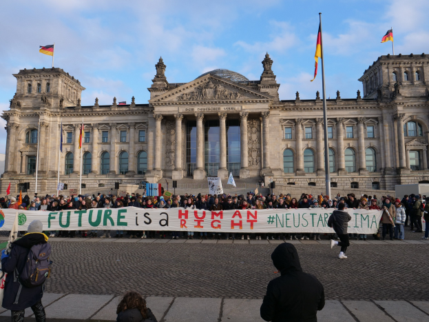 Eine Gruppe von Menschen vor dem Reichstagsgebäude in Berlin mit einer Fahne, auf der der Text 'Zukunft ist ein Mensch Neustar ima' steht und umgeben von Flaggen.