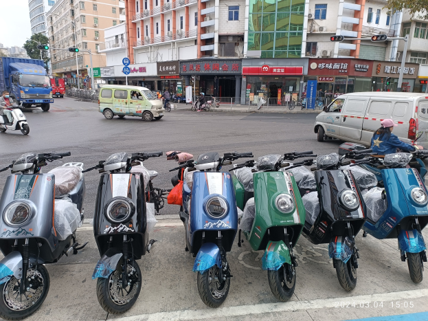 Eine Reihe von Elektro-Scootern, die an einer Straße geparkt sind, mit einer Person in einer Mütze davor, vor dem Hintergrund von Gebäuden, Ampeln, Bäumen und einem klaren Himmel.