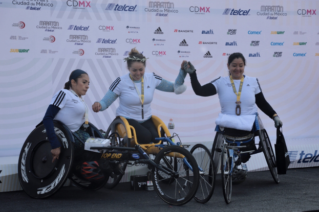 Drei Frauen im Rollstuhl auf einer Rennbahn, mit Medaillen und einem Banner im Hintergrund.
