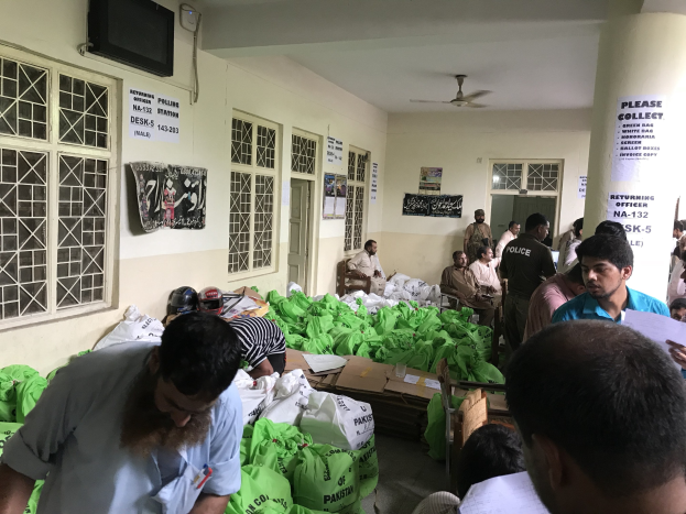 Menschen versammelten sich in einem Raum mit Lebensmittelbeuteln, einige saßen auf Stühlen und andere standen, einige hielten Papiere, mit Fenstern, Plakaten, einem Fernseher, Türen, einer Säule und Deckenventilatoren.