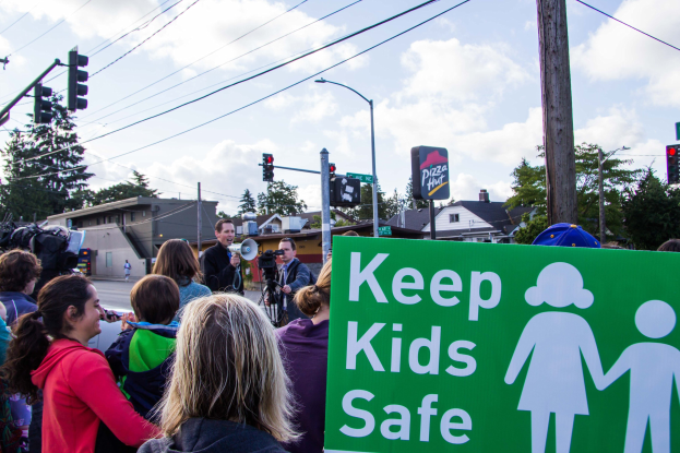 Gruppe von Menschen an einer Straßenkreuzung mit einem "Kinder sicher"-Schild, mit einem Mann mit Megafon im Vordergrund und Gebäuden, Bäumen und Verkehrsinfrastruktur im Hintergrund.