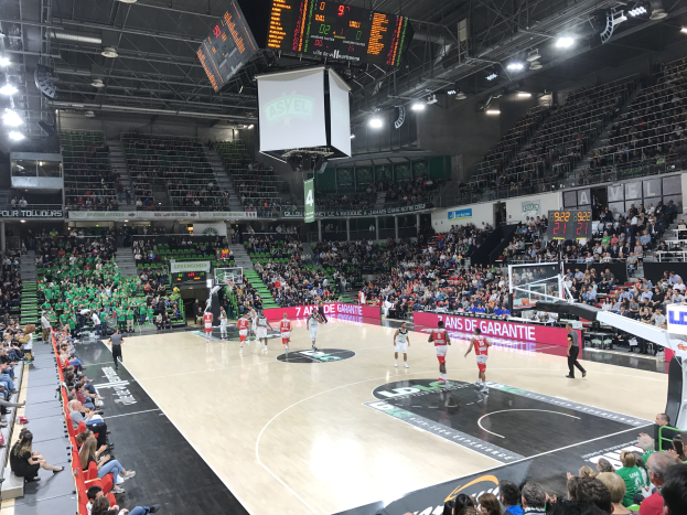 Ein Basketballspiel in einer großen Arena mit Zuschauern auf den Tribünen, Anzeigetafeln mit "Asvel vs Galatasaray" und einer Decke mit Lampen und einem Bildschirm.