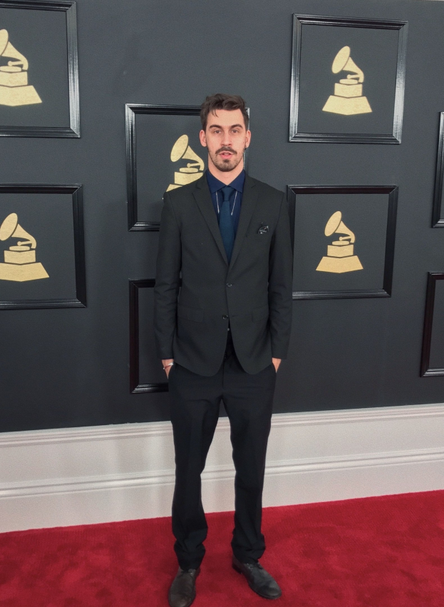 Ein Mann in einem Anzug steht auf dem roten Teppich bei den 55. Annual Grammy Awards, umgeben von einer Wand, die mit Bildern geschmückt ist.