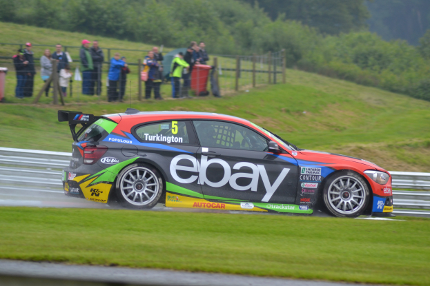Ein Auto, das auf einer Rennstrecke fährt und Rauch ausstößt, umgeben von Gras, einem Zaun, Mülltonnen, Menschen, Bäumen und einem klaren blauen Himmel.