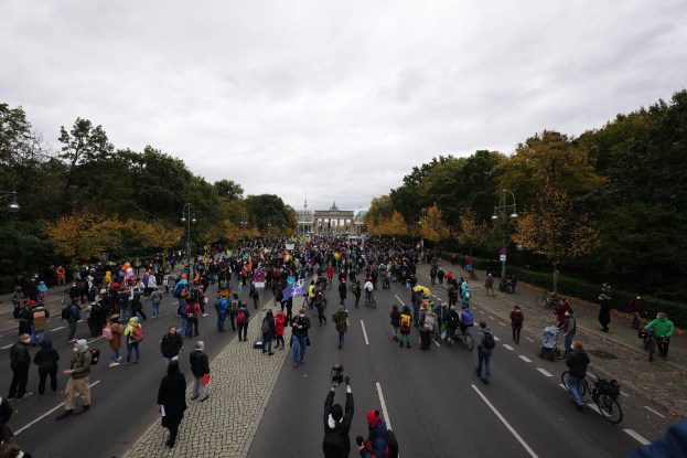 Eine große Gruppe von Menschen marschiert auf einer von Bäumen gesäumten Straße in Berlin, einige halten Kameras, mit einem Gebäude und einem klaren Himmel im Hintergrund.