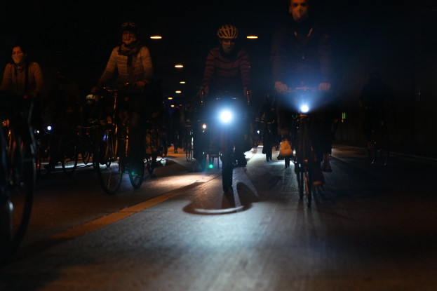 A group of people wearing helmets ride bicycles down a city street at night, illuminated by city lights.