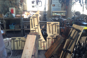 Eine Werkstatt voller verschiedener Werkzeuge, Geräte und Holzplanken, mit einem Stuhl links, Werkzeugen an der Wand und einem Fenster im Hintergrund.