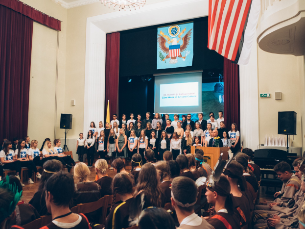 Eine Gruppe von Menschen steht vor einem sitzenden Publikum auf einem Podium mit Mikrofon, flankiert von Fahnen und Lautsprechern, während eines 2. Kunst- und Kulturwochen-Events, mit einem Bildschirm, Vorhängen und einem Kronleuchter im Hintergrund.