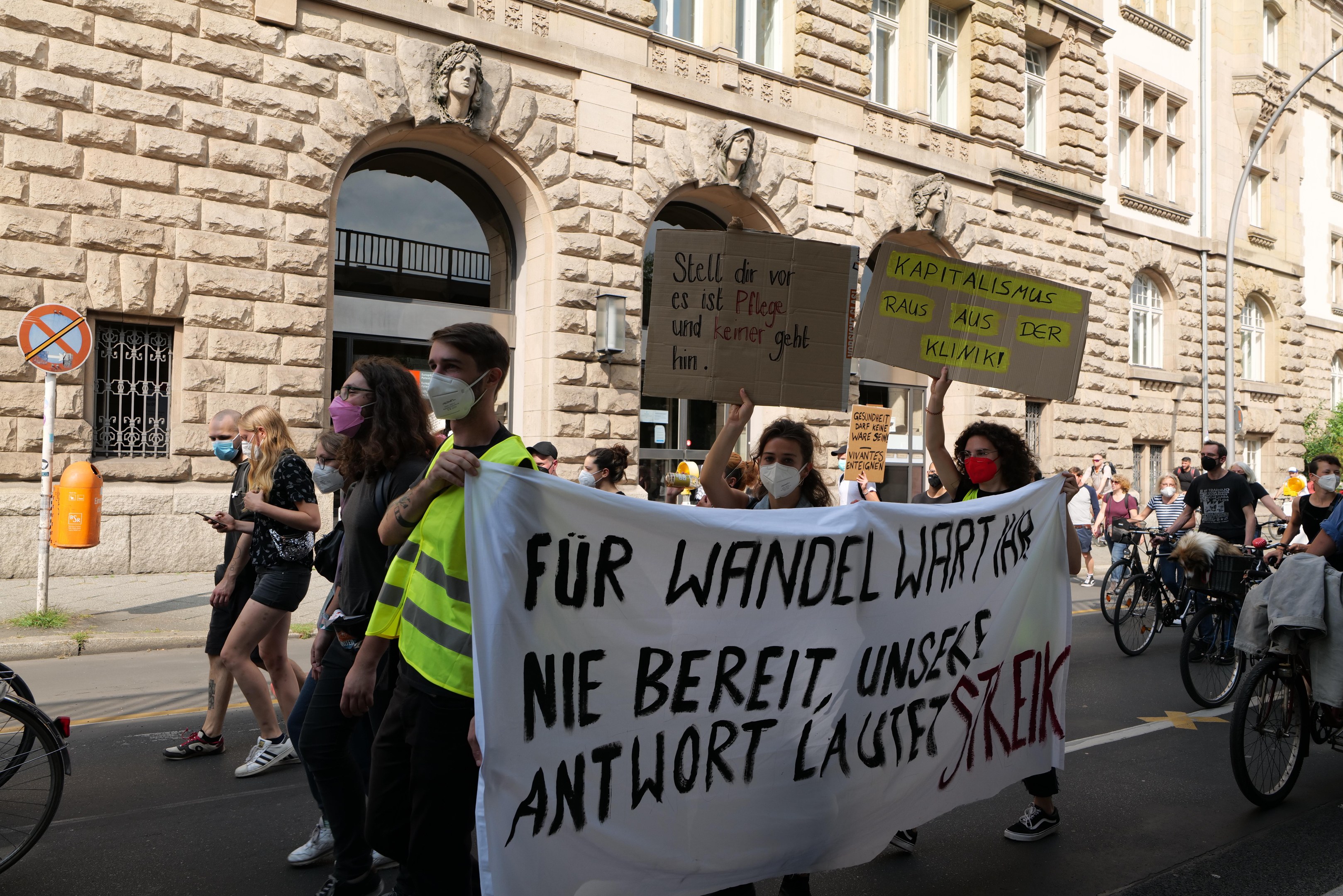 Eine Gruppe von Menschen marschiert durch eine Straße, einige halten Protestschilder und andere fahren Fahrräder, mit einem historischen Gebäude im Hintergrund, das Fenster, Bögen, Säulen und Skulpturen aufweist.