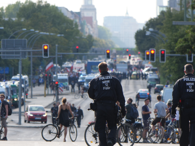Zwei Polizeibeamte neben einer Gruppe von Radfahrern auf einer Straße mit Fahrzeugen, Verkehrszeichen, Bäumen, Gebäuden und einem klaren Himmel.
