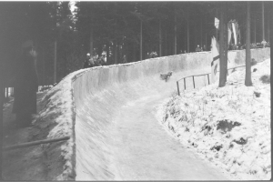 Ein Schwarz-Weiß-Foto eines Menschen, der auf einem Bobschlitten eine schneebedeckte Anhöhe hinunterfährt, umgeben von einem Zaun und Bäumen, mit einigen Menschen im Hintergrund.