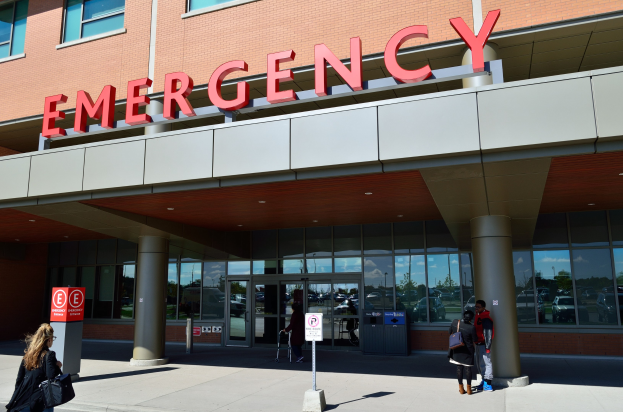 Eine Gruppe von Menschen geht vor einem Notfallgebäude mit Glaswänden, Säulen, einem Namensschild, Hinweistafeln auf Pfählen und einem Mülleimer.