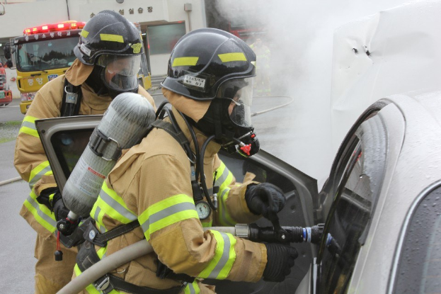 Feuerwehrleute in Schutzausrüstung löschen ein brennendes Auto auf einer Straße mit Fahrzeugen und einem Gebäude im Hintergrund, Rauch steigt aus dem Fahrzeug.