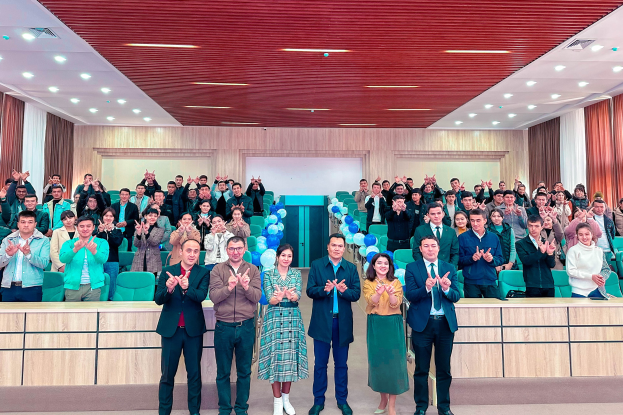 Eine Gruppe von Menschen steht vor einem Publikum, einige klatschen, in einem Raum mit Stühlen, Tischen, Luftballons, Vorhängen und Deckenleuchten, bei der Eröffnungsfeier einer neuen Schule.