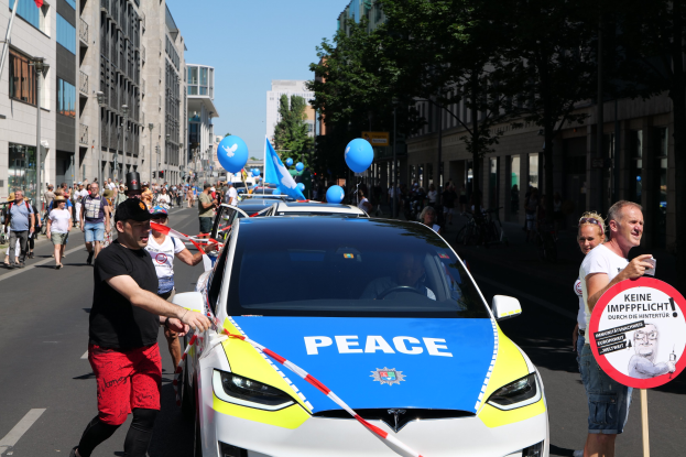 Eine Gruppe von Menschen geht neben einem Polizeiwagen her, der mit blauen, gelben und roten Schleifen geschmückt ist und auf dem das Wort "Frieden" zu sehen ist, mit Gebäuden, Bäumen und Fahnen im Hintergrund unter einem klaren blauen Himmel.