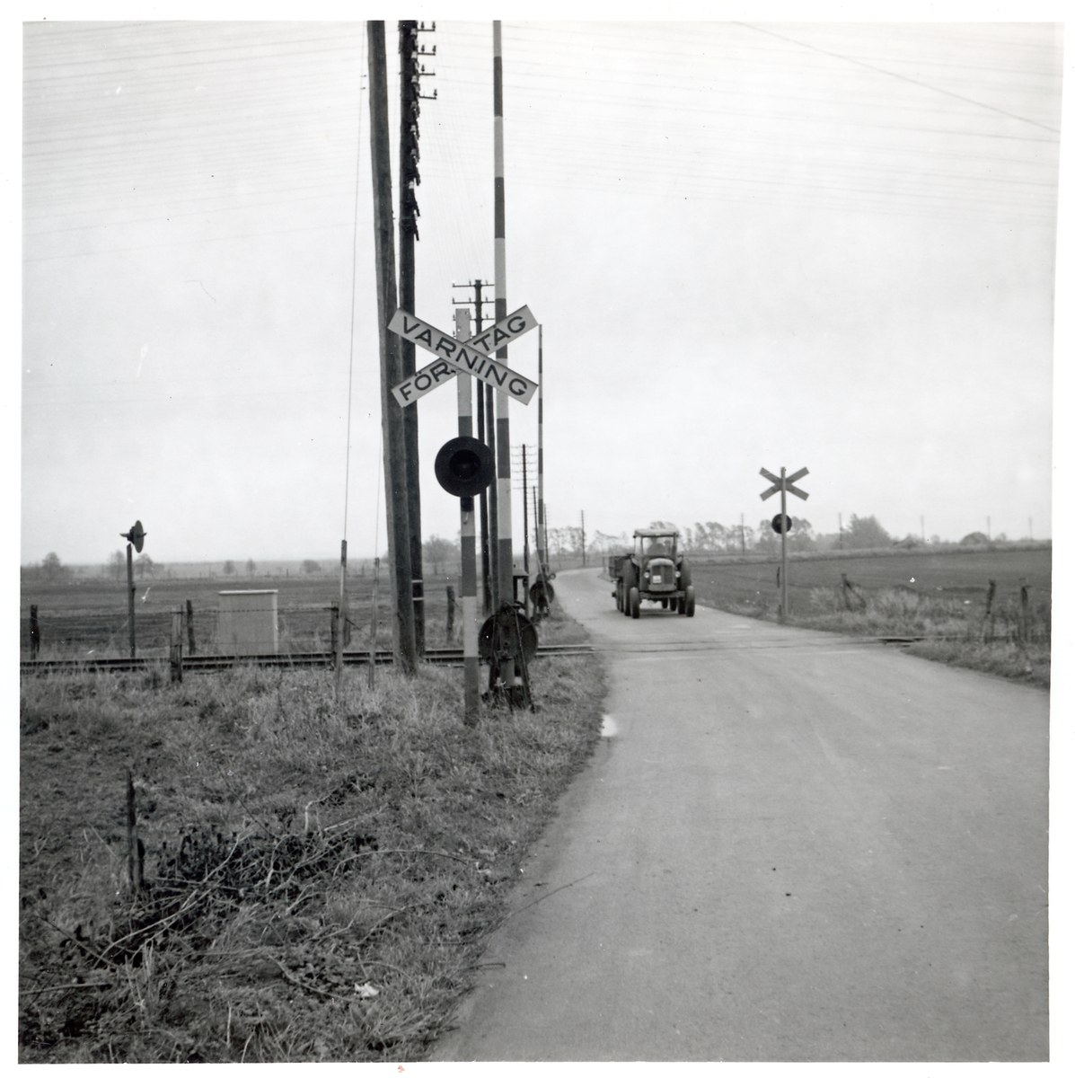 Schwarß-weißes Foto eines Zuges, der eine Straße mit einem Fahrzeug auf der Seite und von Pfosten mit Schildern, Bäumen und einem klaren Himmel im Hintergrund quert.