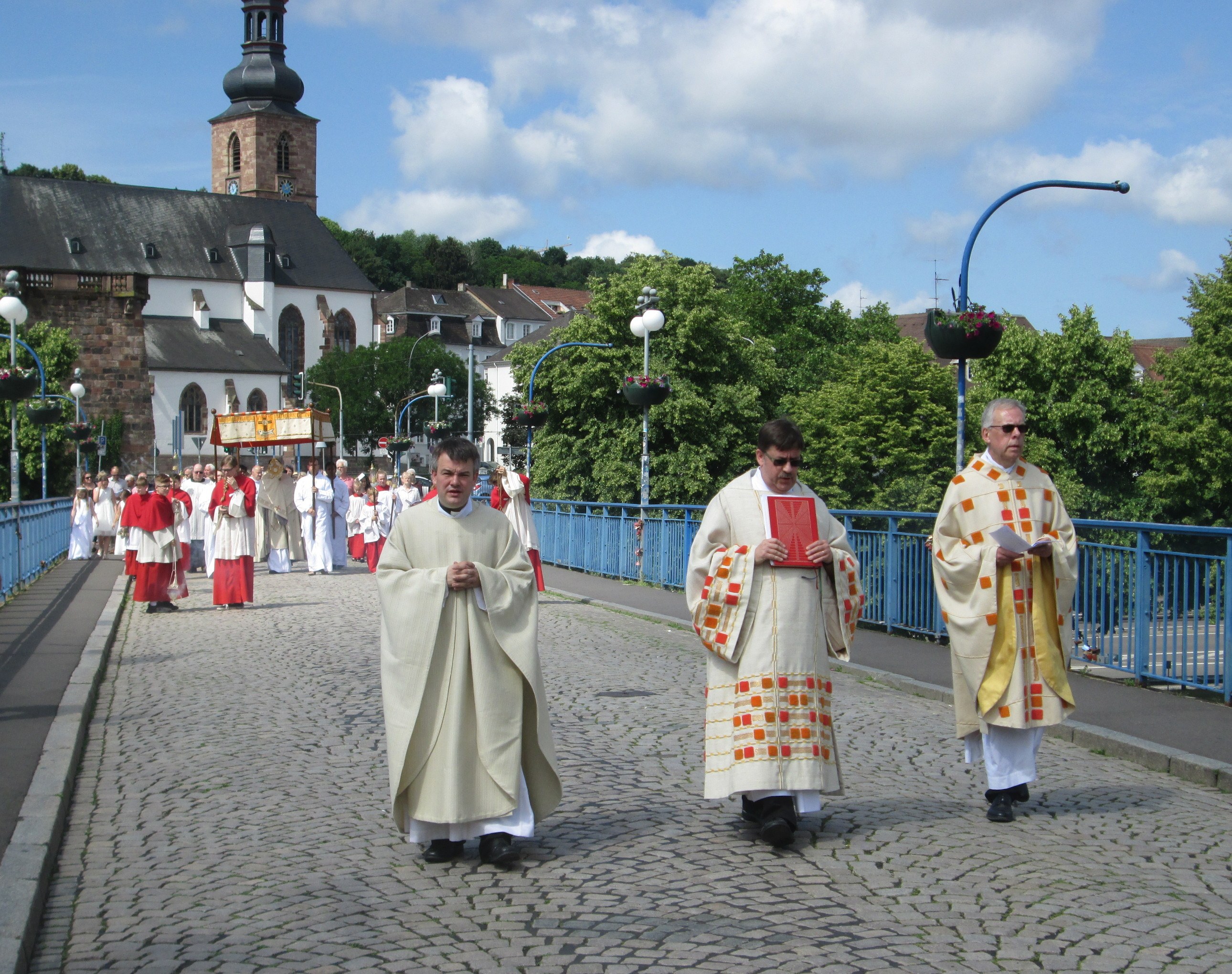Eine Gruppe von Priestern, die eine Straße mit Laternen, Geländern und Bäumen entlanggehen; im Hintergrund sind Gebäude und ein bewölkter Himmel zu sehen; einer der Priester hält ein Buch.