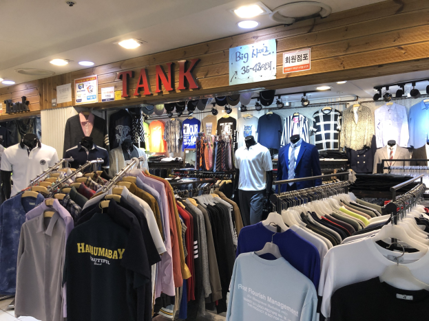 Ein modernes Bekleidungsgeschäft mit zahlreichen Shirts, die auf Gestellen und von Schaufensterpuppen in aktuellen Outfits getragen werden, mit beleuchteter Decke und informativen Wandtafeln.