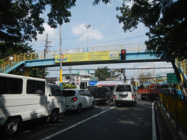 Vielbefahrene Straße mit Fahrzeugen, eine Brücke mit Geländern und Treppen, Laternen, Verkehrsampeln, Informationsschilder, Bäume, Gebäude und ein bewölkter Himmel.