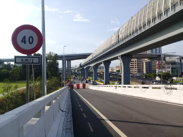 Straße mit Tempolimit-Schild, Fahrzeuge, Brücke mit Pfeilern, Laternen, Bäume, Gebäude und bewölkter Himmel im Hintergrund.