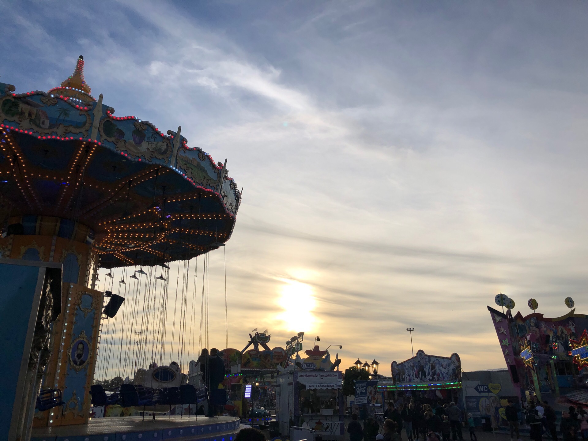 Ein Karussell auf einem Rummelplatz mit Menschen darauf, beleuchtet von der untergehenden Sonne, umgeben von Rummelplatzattraktionen, Pfählen und Lichtern, mit einem bewölkten Himmel im Hintergrund.
