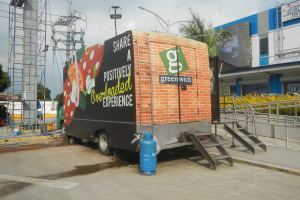 Ein großer Lastwagen parkt vor einem Gebäude mit einer Tafel, auf der "Teilen Sie ein positiv überladenes Erlebnis" steht, mit Stufen, einem Zylinder auf dem Boden, Geländern, Strommasten, Bäumen und einem bewölkten Himmel.