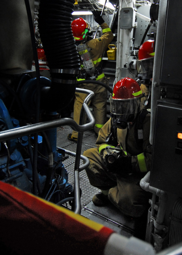 Feuerwehrleute in Helmen und Schutzausrüstung arbeiten an einem Löschwagen mit Rohren und Gegenständen im Hintergrund, unter einem Licht an der Spitze.