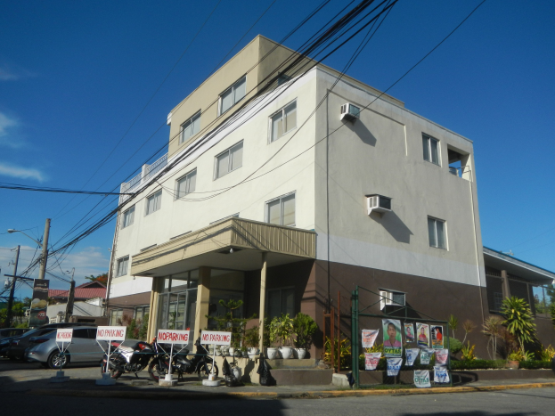 Wohnanlage mit parkenden Autos, Schildern, Versorgungsmasten, Bäumen und einem bewölkten Himmel.