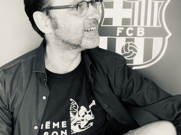Ein Mann mit Brille und schwarzem Hemd lächelt vor einem schwarz-weißen FC Barcelona-Logo.