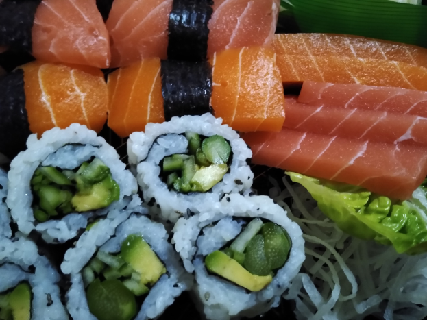 Ein schwarzer Teller mit verschiedenen Sushi-Stücken, darunter Lachs, Avocado und Gurke, umgeben von ein paar Blättern.