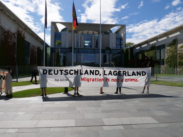 Gruppe von Menschen mit einem Transparent mit der Aufschrift "Deutschland, Lagerland Migration ist kein Verbrechen" vor einem Fenster mit einem Metallzaun, Pflanzen, Bäumen und Fahnenmästen unter einem bewölktem Himmel.