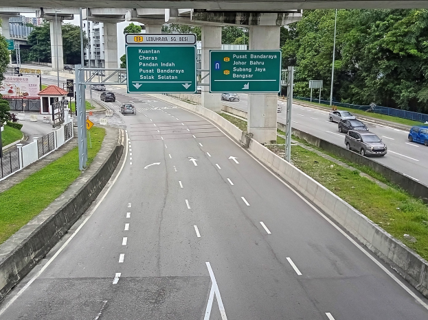 Autobahn mit mehreren Autos, umgeben von Gras, einem Zaun, Strommasten, Holzplanken, Bäumen, Gebäuden und einer Brücke im Hintergrund, mit einem sichtbaren Verkehrszeichen.
