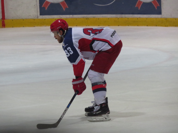 Hockeyspieler in voller Ausrüstung auf dem Eis mit einem Stock, der einer Wand mit Bannern im Hintergrund gegenübersteht.