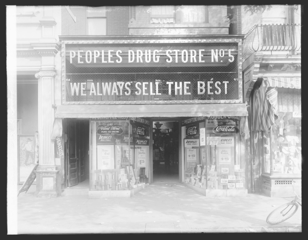 Schwarzes und weißes Foto einer Geschäftsfront mit der Aufschrift "People's Drug Store No. 5" und einem Fahrrad davor sowie verschiedenen Gegenständen im Inneren.