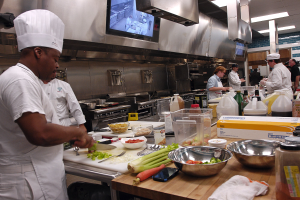 Eine Gruppe von Köchen in einer Küche, die Essen auf einem Schneidbrett zubereiten, wobei ein Koch in der foreground in einer weißen Schürze und einer Mütze steht, ein Messer hält und von verschiedenen Utensilien, Schüsseln, Gemüse, einem Mobiltelefon, Flaschen und anderen Gegenständen umgeben ist, mit einem Fernseher und Deckenlampen im Hintergrund.
