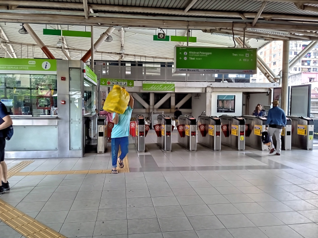 Menschen gehen durch einen Bahnhof mit Metalldetektoren, Hinweistafeln, Metallstangen, Deckenleuchten und Fenstern im Hintergrund.
