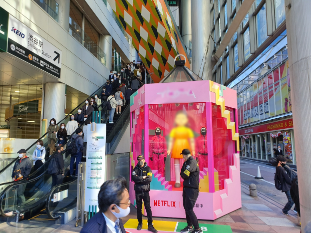 Eine Gruppe von Menschen steht vor einem Gebäude mit Glaswänden, Säulen und Texttafeln, einige tragen Masken, während eine Rolltreppe mit Menschen in der Nähe fährt; eine pinkfarbene Box in der Mitte zeigt Schaufensterpuppen und trägt den Text "Netflix's neuer Netflix-Stand in Tokio."