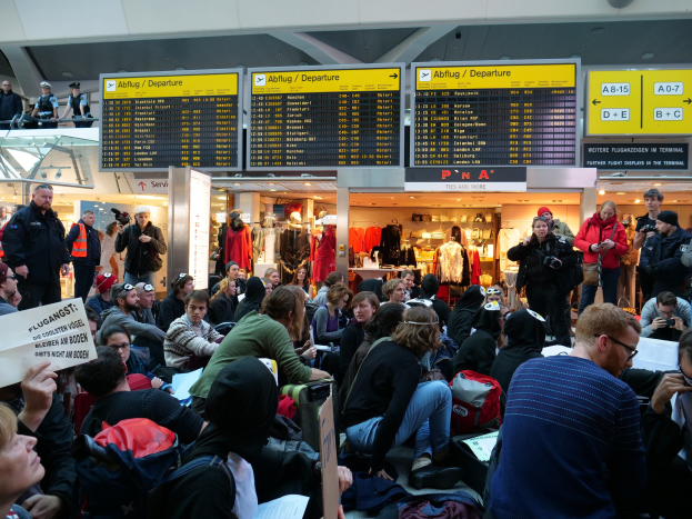 Eine große Gruppe von Menschen sitzt und steht in einem Flughafen während einer Demonstration, wobei einige Taschen und Papiere halten, während Bretter mit Text, Schaufensterpuppen in Kleidern und Deckenleuchten im Hintergrund zu sehen sind.