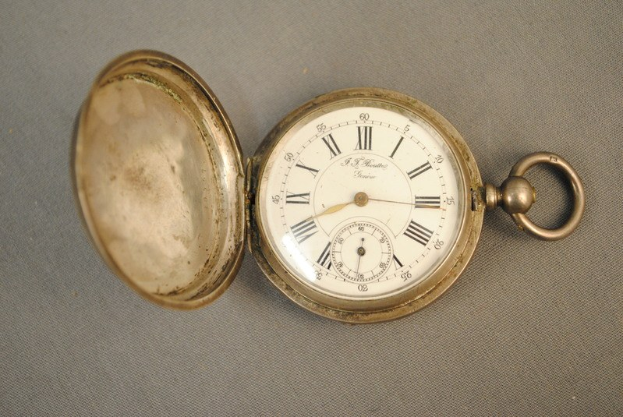 Eine silberne Taschenuhr mit einem weißen Zifferblatt mit römischen Zahlen und einem Deckel, auf einer grauen Oberfläche.