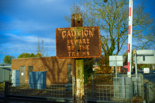 Warnschild an einem Bahnübergang-Zaun mit Bäumen, Strommasten, einem Gebäude, Containern, einer Straße mit Fahrzeugen und einem bewölkten Himmel.