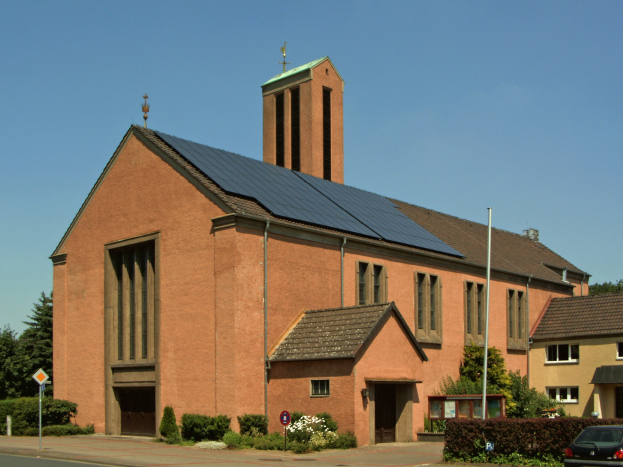 Kirche mit Solarpaneelen auf dem Dach, umgeben von Gebäuden, Straßenlaternen, Straßenlaternen, Kraftfahrzeugen, Verkehrsschildern, Büschen und Bäumen unter einem klaren blauen Himmel.