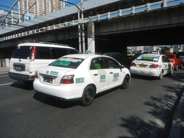 Eine Gruppe von Taxis steht entlang einer Straße mit einer Brücke, Strommasten, Gebäuden, Bäumen und einem klaren blauen Himmel im Hintergrund.